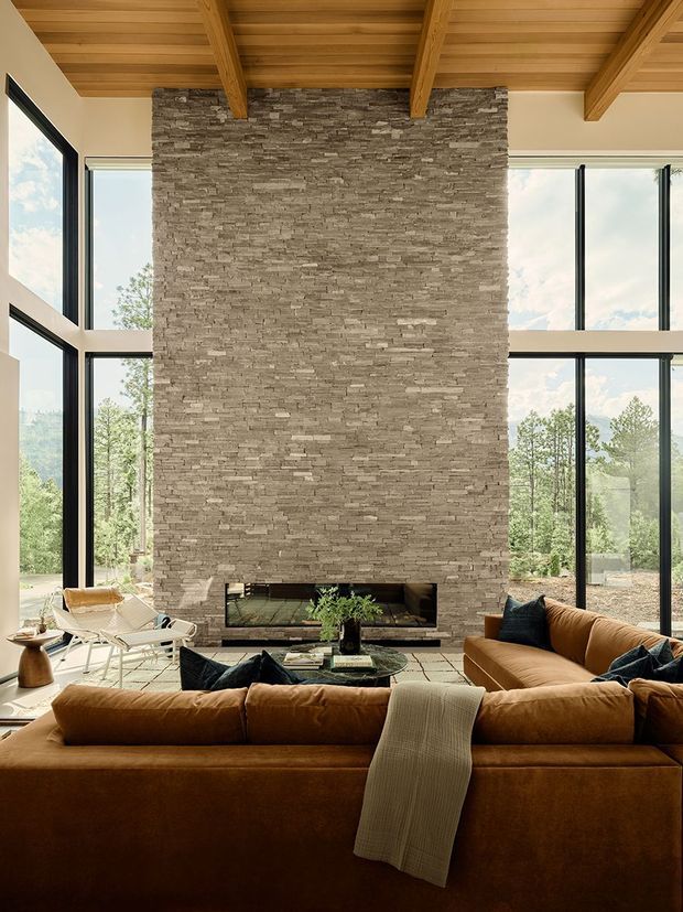 A spacious double-height living room centered on a tall stacked-stone fireplace wall. Warm timber ceiling boards and exposed beams contrast with expansive black-framed floor-to-ceiling glazing overlooking trees. A large cognac-brown sectional sofa faces the fireplace, with lounge chairs and a low coffee table arranged in front. The room has a warm, natural, refined contemporary lodge-like atmosphere.