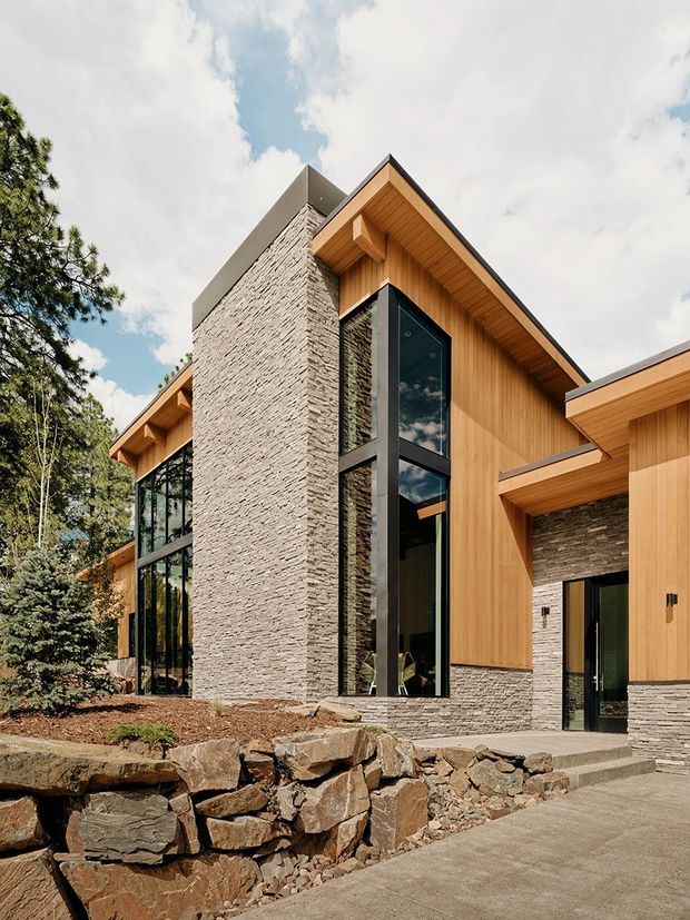 Exterior view of a contemporary multi-volume house set among trees, featuring steep gabled roof forms, warm vertical timber cladding and soffits, dark metal-framed floor-to-ceiling glazing, and prominent textured natural-stone walls and retaining landscaping. The entry is visible at ground level, approached by a concrete path and stone steps.