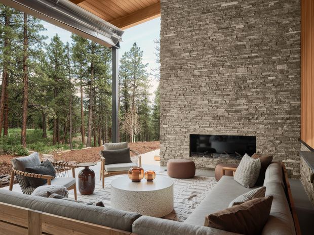 A contemporary indoor-outdoor living room opening onto a pine forest, with a large sectional sofa, lounge chairs, round coffee table, and a prominent stacked natural-stone fireplace wall. The room has a warm timber-lined ceiling, expansive open glazing, neutral furnishings, and a sheltered terrace-like connection to the surrounding landscape.