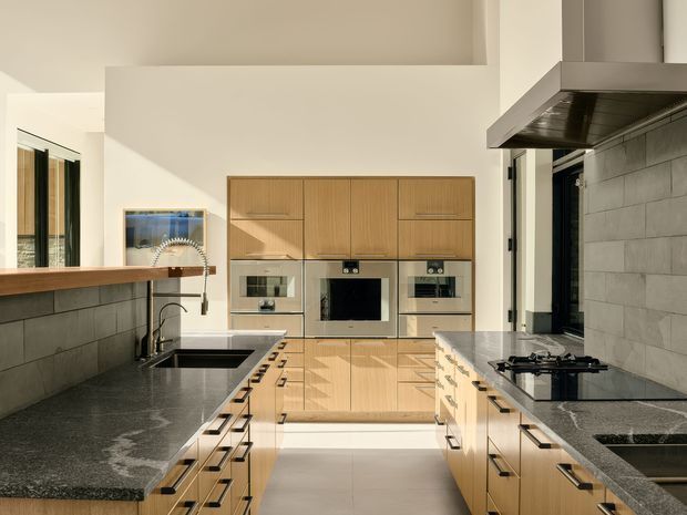 A bright contemporary kitchen viewed from the main preparation area, featuring a large U-shaped layout with dark stone benchtops, light timber cabinetry, integrated ovens and microwave, a gas cooktop, deep sink, tiled splashback, and a substantial stainless-steel rangehood. A timber-topped breakfast bar and open glazing bring natural light into the space.