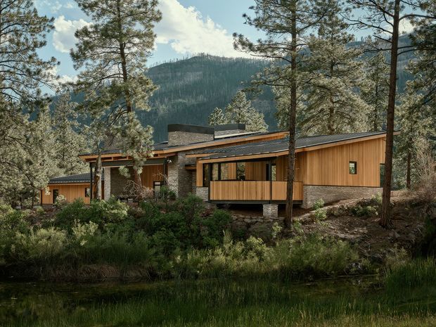 A low-profile contemporary house set among pine trees in a mountainous forest landscape. The building has horizontal timber cladding, dark stone or masonry walls, broad roof overhangs, extensive glazing, and a shallow pitched or near-flat roofline. A pond or wetland edge is visible in the foreground, with a forested mountain behind.