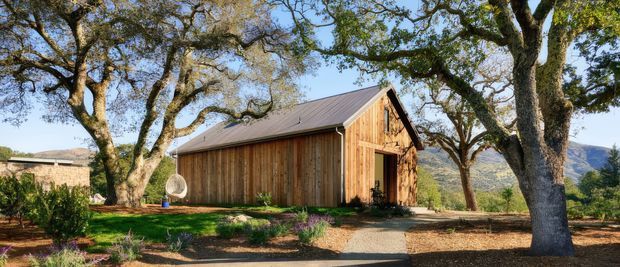 Exterior view of a rustic timber-clad barn-like building set within a landscaped rural site. The long single-storey structure has a steep gabled roof with dark metal roofing, vertical natural timber cladding, and large glazed openings at the gable end. Mature spreading trees frame the building, with gravel paths and planted garden beds in the foreground and hills visible beyond.
