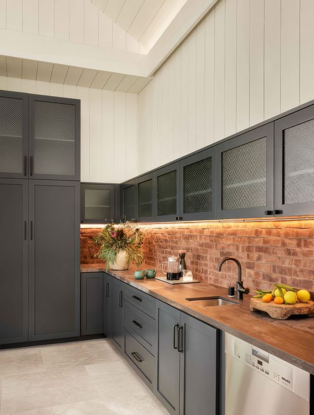 A compact, carefully detailed kitchen with dark grey shaker-style cabinetry, textured glass-front upper cupboards, a warm timber benchtop, and an exposed red brick splashback illuminated by under-cabinet lighting. White vertical wall panelling and a pitched, panelled ceiling brighten the room, while a stainless sink, black tapware, integrated dishwasher, and pale tiled floor complete the fit-out.