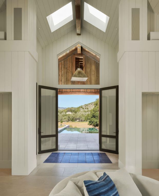 A bright, double-height entry hall with white vertical wall panelling and a steeply pitched, gabled ceiling. Two large skylights and a central exposed timber beam illuminate the space. Open dark-framed glazed double doors lead to a tiled terrace, swimming pool and landscaped rural hillside beyond. A rustic timber-lined gable feature and pendant light add warmth, while a partially visible upholstered seating piece sits in the foreground.
