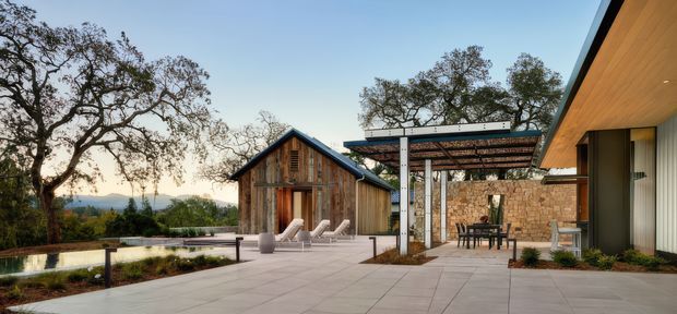 A contemporary outdoor terrace and pool area overlooking a wooded landscape. The scene includes a timber-clad gabled pavilion, broad pale paving, lounge chairs beside a long pool, and a covered outdoor dining area with a pergola and stone wall. A modern building with warm timber soffit and large glazing frames the right side.