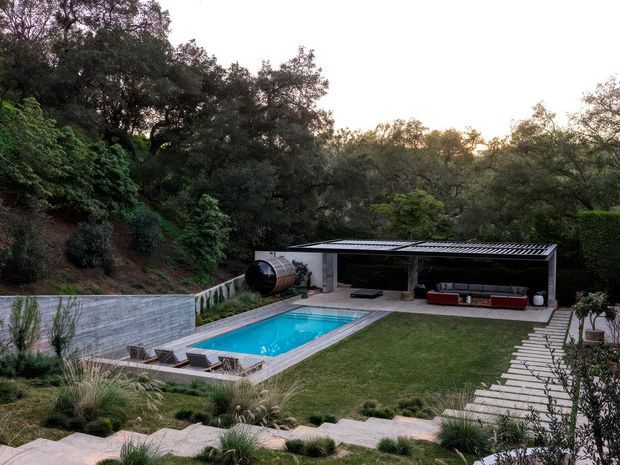 Elevated view of a landscaped residential outdoor area with a rectangular swimming pool, paved pool surround, lawn, stepped stone paths, dense mature vegetation, and a covered open-sided pavilion containing outdoor seating. A corrugated metal roof and concrete walls define the pavilion, while a curved retaining wall borders the pool.