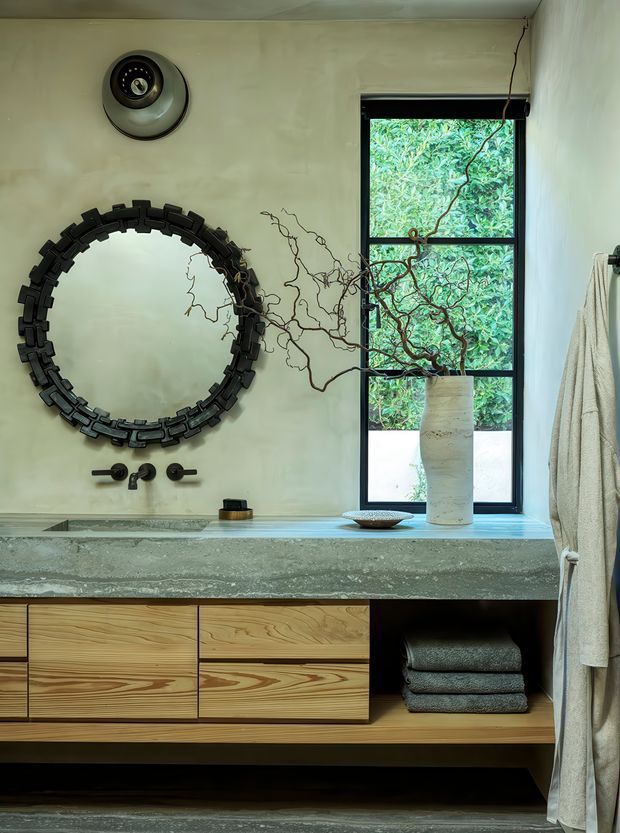 A refined bathroom vanity with a wall-mounted round mirror in a dark geometric frame, black wall-mounted tapware, a textured concrete or stone benchtop, and warm natural timber drawers. A tall black-framed window overlooks dense greenery, while a sculptural vase with branches and folded towels add organic detail. The room has a calm, earthy, contemporary feel with a mix of raw textures and natural materials.