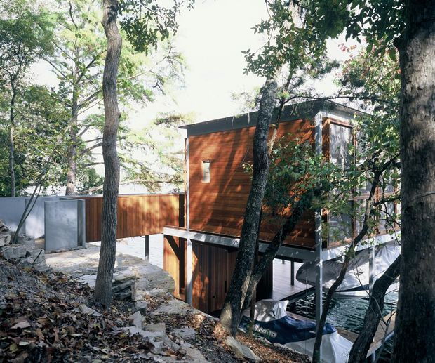 A modern elevated timber-clad house nestled among mature trees on a sloping, wooded site. The compact multi-level structure features a dark flat roof, warm brown vertical timber cladding, extensive glazing, and a raised deck or balcony with metal supports. Rocky ground, tree trunks, and a partially visible lower-level outdoor area reinforce the bush setting.