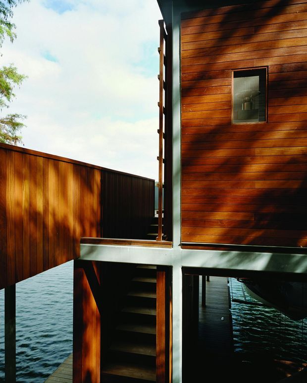 Exterior architectural view of a contemporary elevated structure over water. The building features rich horizontal timber wall cladding, dark metal structural framing, an external timber stair, and a timber privacy screen or adjoining wall. Water is visible beneath and beside the raised building, emphasizing its stilted or waterfront construction.