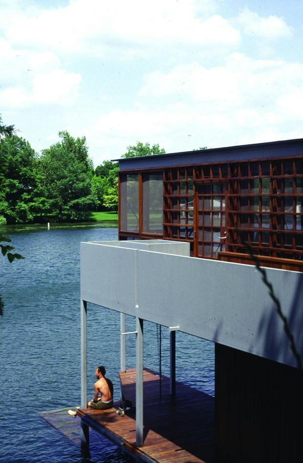 Exterior view of a modern multi-level building projecting over a lake or pond. The structure features a flat roof, extensive glazing with a distinctive timber lattice screen, smooth light-grey wall planes, slender supports, and a timber waterside deck with a small dock. Mature trees and water surround the building.