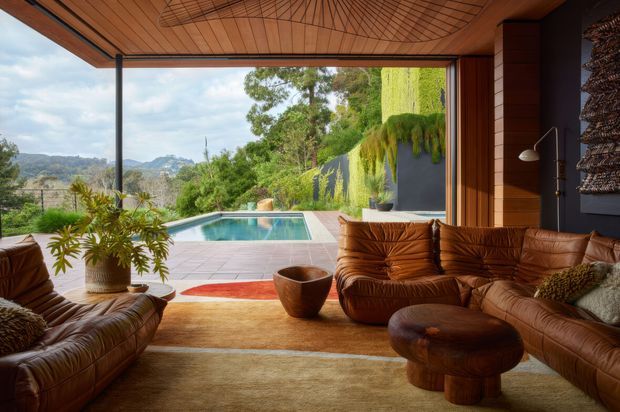 A warm mid-century-inspired living room opening directly onto a swimming pool and landscaped outlook. The room features extensive timber wall and ceiling lining, dark feature walls, large open glazing, low brown leather sofas, a rounded timber coffee table, and a muted layered rug. The pool terrace and green hillside are visible beyond the open facade.