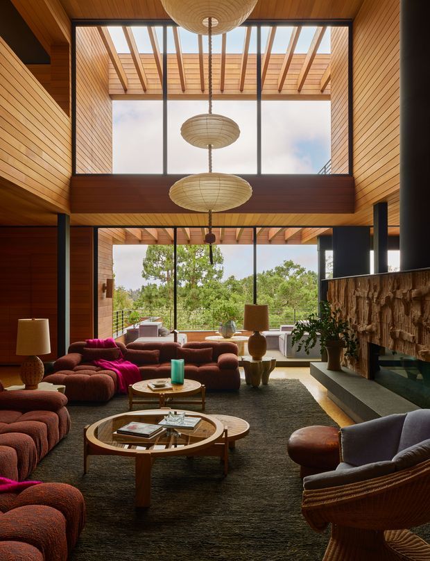 A spacious, richly timber-lined living room with a dramatic double-height glazed wall and open roof pergola above. Multiple sculptural woven pendant lights hang over a warm mid-century-inspired seating arrangement with deep rust sofas, patterned cushions, rounded timber coffee tables, and a dark textured rug. A large stone or textured feature fireplace anchors the right side, while full-height glazing opens views toward leafy landscaping.