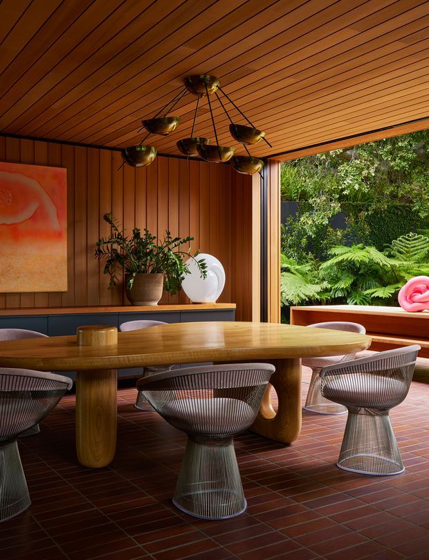 A richly detailed dining room with a large rounded timber dining table and sculptural wire-frame chairs. The room features warm vertical timber wall panelling, a slatted timber ceiling, red-brown brick flooring, dark built-in cabinetry, a suspended sculptural brass-toned light fixture, and artwork and plants. Full-height opening to a lush fern-filled garden creates a strong indoor-outdoor connection.