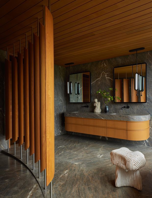 A luxurious, moody bathroom with a long floating double vanity, integrated curved timber cabinetry, dark veined stone benchtop and matching stone wall and floor surfaces. Two mirrors with sculptural pendant lights hang above the vanity. A dramatic curved timber slat partition screens the bathroom entrance, while a small textured upholstered stool sits in the foreground.