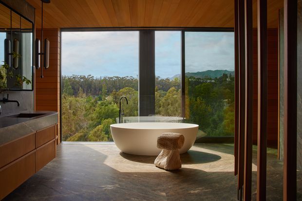 A warm, contemporary bathroom with a freestanding white oval bathtub positioned in front of expansive floor-to-ceiling windows overlooking a wooded landscape and hills. The room features rich timber wall and ceiling lining, dark stone-like flooring, timber vanity cabinetry, a large mirror, black plumbing fixtures, and a sculptural wooden stool. A partially open timber-framed doorway is visible at right.