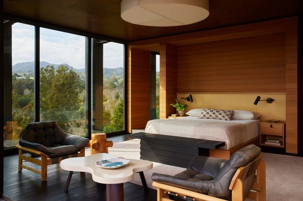 A warm, wood-lined bedroom with a large bed set against a horizontal timber feature wall and integrated bedside shelving. Floor-to-ceiling glazing provides expansive views of a wooded, mountainous landscape. Two leather lounge chairs and a sculptural coffee table create a sitting area in the foreground. The room has dark timber flooring, a large circular ceiling light, and a refined mid-century-inspired atmosphere.