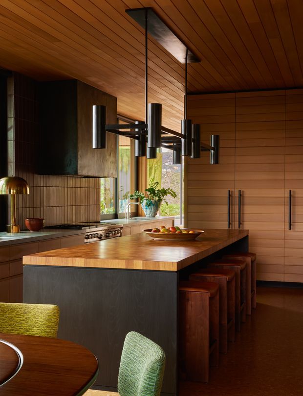 A richly detailed contemporary kitchen with extensive warm timber panelling on the ceiling and walls, dark cabinetry, a large timber-topped island with integrated seating, and sculptural black cylindrical pendant lights. A tiled backsplash, stainless-steel range, sink, greenery, and dining furniture are also visible. The space has a warm, moody, refined character with strong natural-material detailing.