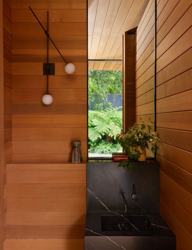 A compact, warmly lit bathroom clad extensively in horizontal timber wall and ceiling panels. A black stone vanity with integrated basin and wall-mounted dark metal tapware sits beneath a tall mirror, with a narrow window opening to lush greenery. Minimal spherical pendant lights and a small vase of foliage add understated natural detail.