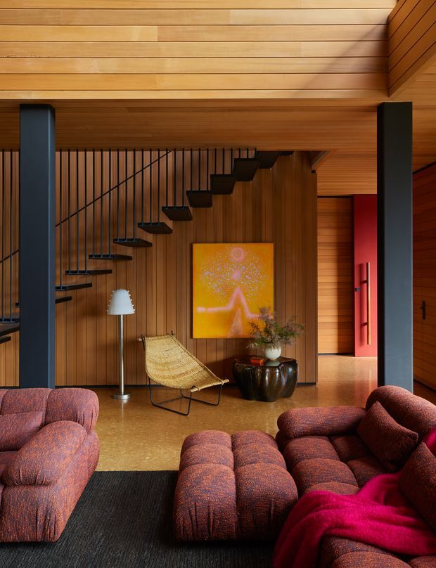 A warm, richly detailed living room with extensive timber wall and ceiling lining, a sculptural open staircase, dark structural columns, and deep red textured modular seating arranged around a black rug. A woven lounge chair, floor lamp, artwork, ceramic planter, and a vivid red doorway add visual interest. The space has a strong mid-century-inspired architectural character with contemporary furnishings and dramatic warm lighting.