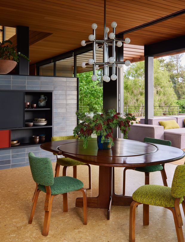 A stylish open-plan dining and living area with a large round polished timber dining table, upholstered mid-century-inspired chairs in green and yellow, and a sculptural chrome-and-globe pendant light. The room features a warm timber slatted ceiling, dark structural beams and cabinetry, a grey tiled feature wall with open shelving, and extensive glazing opening to leafy outdoor views. A lounge area is visible beyond the dining space.