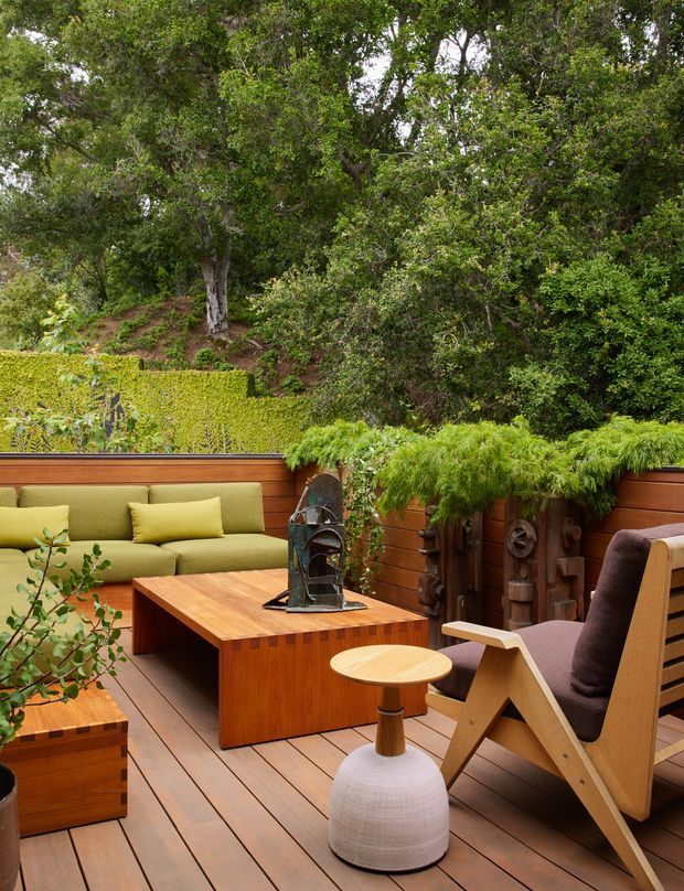 A lush, enclosed outdoor lounge on a timber deck, furnished with a built-in olive-green sectional sofa, a large timber coffee table, and a sculptural timber-framed armchair. Wooden privacy screens, raised planters, and dense greenery surround the space, creating a sheltered garden setting. The overall aesthetic is warm, natural, and mid-century influenced.
