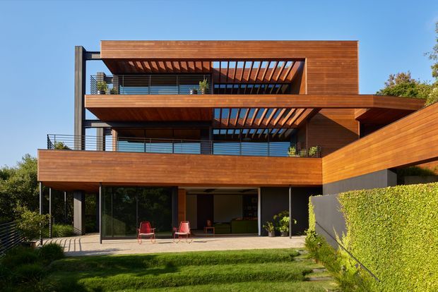 A contemporary multi-storey residential house viewed from the rear garden. The building features extensive horizontal timber cladding, dark structural framing, large glazed openings, projecting balconies, and layered pergola-like timber sun-shading over outdoor terraces. A covered ground-level patio opens to the lawn, with a landscaped hedge and side access path.