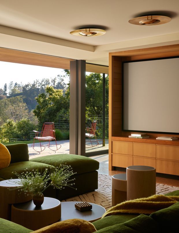 Warm contemporary living room with a large wall-mounted television set into a timber feature wall and integrated timber cabinetry. Floor-to-ceiling glazing opens onto a terrace with garden views, while sculptural ceiling lights, rounded coffee tables, deep green seating, and textured furnishings create a refined mid-century-inspired interior.