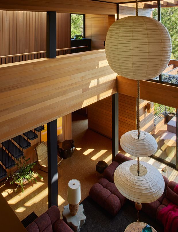 A warm, wood-lined double-height living room viewed from an upper-level landing. The space features extensive horizontal and vertical timber panelling, black structural posts, large windows, a mezzanine-like upper walkway, and multiple oversized spherical paper pendant lights suspended above deep burgundy seating. A staircase, indoor planting, and lower-level lounge area are visible, creating a dramatic and open multilevel interior.