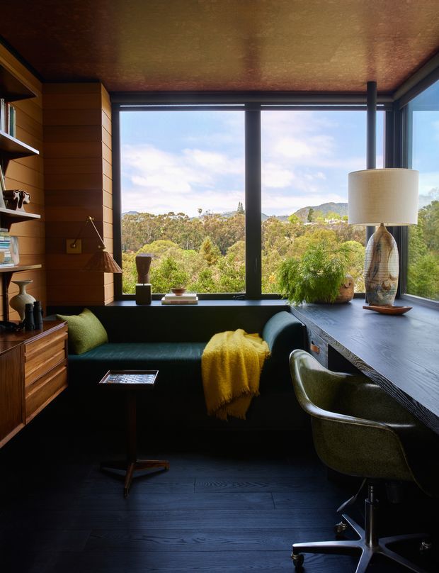 A compact, richly detailed home office or study with a dark built-in window seat, integrated timber shelving and cabinetry, a large corner desk, and a rolling upholstered office chair. Floor-to-ceiling corner glazing overlooks a leafy wooded landscape and hills. The room features dark timber flooring, warm timber wall panelling, a textured metallic ceiling, a sculptural lamp, and green and mustard textiles, creating a moody yet inviting atmosphere.