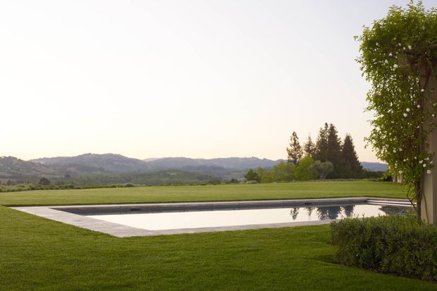 A landscaped outdoor setting with a long rectangular reflecting pool or swimming pool set into a broad, neatly maintained lawn. A clipped hedge and mature trees frame the right edge, while open fields and low rolling hills extend into the distance. The scene has a calm, spacious, resort-like character.