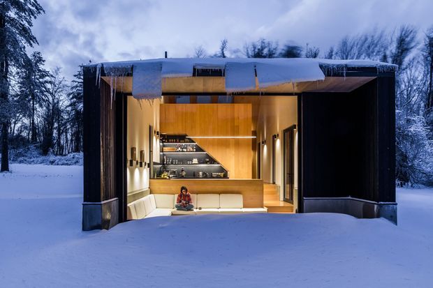 A compact modern cabin-like retreat set in a snowy forest at dusk or night. The front facade is opened fully to reveal a warm timber-lined interior with a built-in cream sectional sofa, a compact lounge area, sculptural dark shelving and cabinetry, and integrated lighting. Snow and icicles cover the flat roof and surround the structure, creating a strong contrast between the cool blue exterior and warm amber interior.