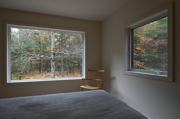 A quiet, compact bedroom with a low-profile bed covered in a dark grey quilt and a simple wooden spindle-back chair in the corner. Two large framed windows provide views into a wooded landscape with autumn foliage. The room has plain warm-grey walls, a white ceiling, minimal furnishings, and abundant natural outlook, creating a calm, understated retreat.