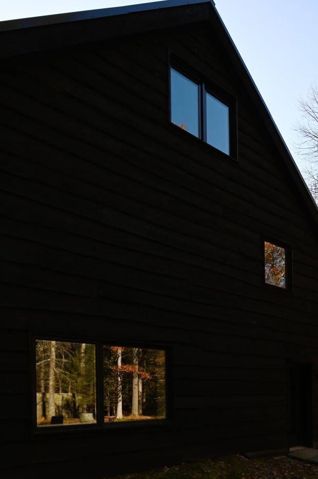 Dark, minimalist two-storey gable-roof house exterior clad in horizontal dark timber weatherboards. Several black-framed windows reflect surrounding woodland, and the steeply pitched roof and simple rectangular form dominate the image. The low-angle view emphasizes the tall facade and deep shadowing.