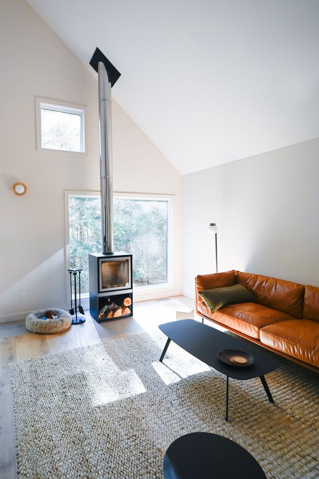 Bright, minimalist living room with a high sloping ceiling, pale walls, large windows, and a freestanding black wood-burning stove with a prominent metal flue. A warm cognac leather sofa, black oval coffee tables, textured neutral rug, floor lamp, and pet bed furnish the space. Natural light and views of trees create an airy, contemporary atmosphere.