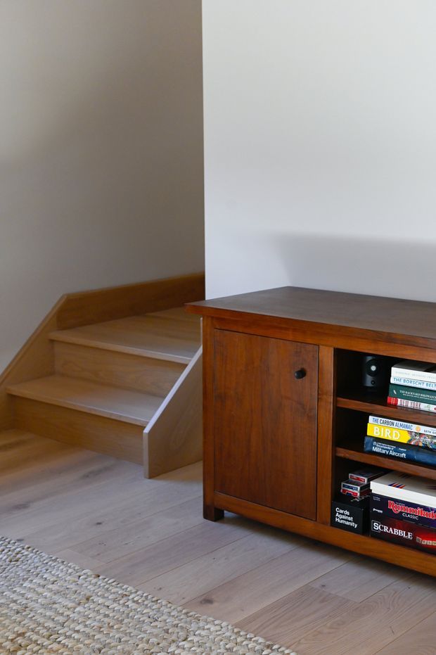 Interior detail showing a compact timber staircase beside a mid-century-style wooden sideboard or media cabinet. The room has pale timber flooring, plain light walls, and a woven natural-fibre rug. The cabinet contains books and board games, while the staircase has open risers and a simple timber balustrade-free design.