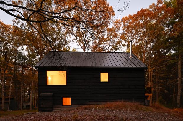 A small, dark-clad cabin set within a wooded landscape during autumn. The compact single-storey building has horizontal black timber-look cladding, a simple dark standing-seam pitched roof, and several warm glowing windows. A chimney projects from the roof, while fallen leaves and mature trees surround the structure, creating a secluded woodland setting.