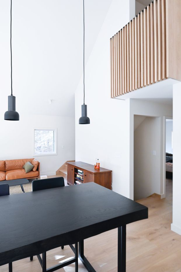 Bright open-plan interior showing a dining area in the foreground with a large black dining table and chairs, opening into a living room with a tan leather sofa and timber media console. White walls, pale timber flooring, black pendant lights, and a timber vertical-slat balustrade overlook the space, suggesting a high or double-height volume.