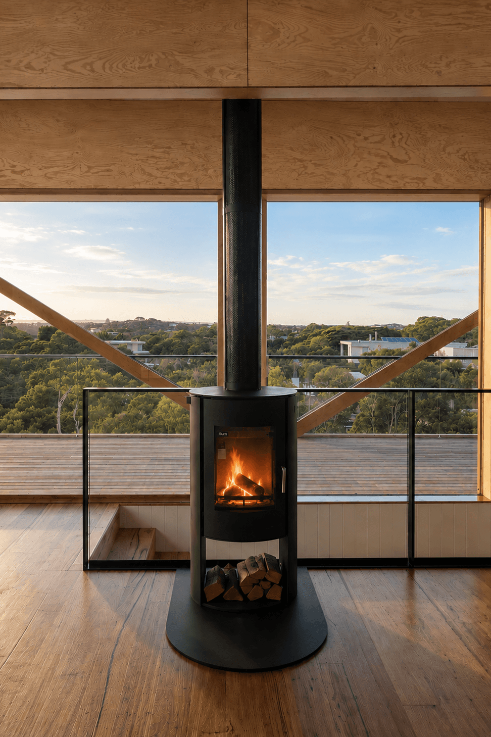 A freestanding Nectre N65 wood stove anchors the living space, framed by expansive glazing that exposes structural timber bracing and coastal treetop view