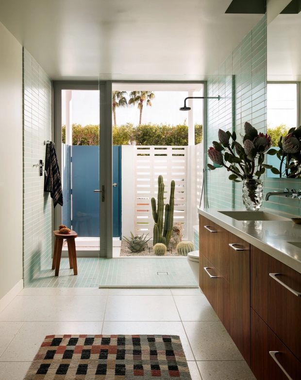 Bright, spacious bathroom with a walk-in shower opening to a planted outdoor courtyard. The room features pale aqua-green horizontal wall tiles, matching small-format shower floor tiles, large light floor tiles, a wood-grain floating vanity with a pale stone countertop, dark metal-framed glazing and door, and a large mirror. A patterned geometric rug and a vase of flowers add decorative colour. The open shower, outdoor screening, cactus planting, and palm trees create a modern mid-century-influenced indoor-outdoor feel.
