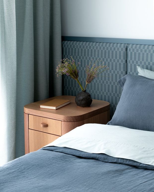 A close-up of a refined bedroom corner featuring a neatly made bed with blue-grey bedding, a blue-grey upholstered headboard with a subtle geometric pattern, and a rounded timber bedside table with drawers. A dark ceramic vase with delicate branches and a book sit on the table, while full-height blue-grey curtains frame the scene. The palette is calm, cool, and muted with warm timber contrast.
