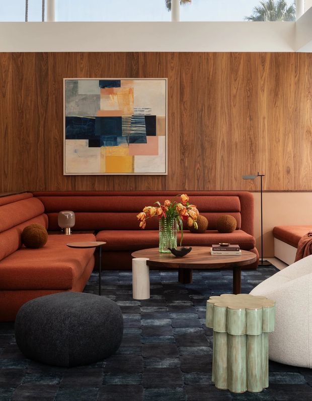 A richly layered living room with a large rust-red sectional sofa arranged around a low oval coffee table. The room features a full-height walnut timber wall, dark blue-grey patterned carpet, sculptural side tables and ottomans, a floor lamp, colourful abstract artwork, and floral arrangements. A high clerestory-like opening brings in daylight and reveals palm trees outside. The overall composition is warm, sophisticated, and highly curated with mid-century-inspired forms and contemporary furnishings.