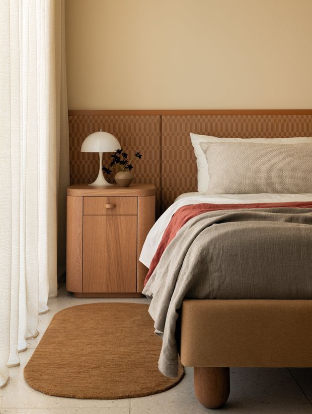 Warm, refined bedroom vignette featuring a low upholstered bed with rounded timber legs, layered neutral and rust bedding, a textured woven headboard, and a curved timber bedside cabinet. A sculptural white dome lamp, small vase, sheer curtains, and a rounded brown rug complete the softly lit interior.