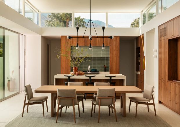 A bright, double-height open-plan dining and kitchen interior with a long timber dining table and upholstered chairs in the foreground. The kitchen features a central island with seating, warm timber cabinetry, a green tiled splashback, and pendant lights. Floor-to-ceiling glazing and high clerestory windows bring in abundant daylight, with palms and a mountain visible outside. A large built-in timber media or storage wall occupies the right side.