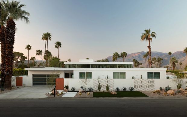 Front exterior view of a low-slung modernist residence in a dry, palm-lined setting. The white single-storey facade has a broad flat roof, horizontal composition, clerestory-like upper glazing, a recessed entry, integrated garage, decorative breeze-block screens, and a landscaped gravel-and-succulent frontage. Mountainous terrain is visible behind the property.