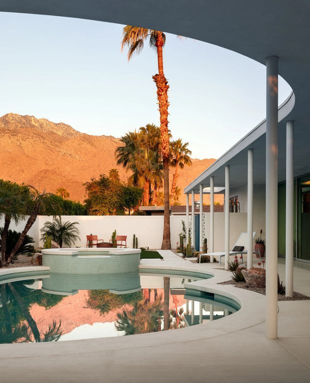 A modernist courtyard pool area beside a low-slung white building, framed by slender round columns and a broad curved roof canopy. The pool has rounded geometric forms and pale mosaic tiling, with desert landscaping, palms and cacti, and dramatic sunlit mountains beyond the courtyard wall.