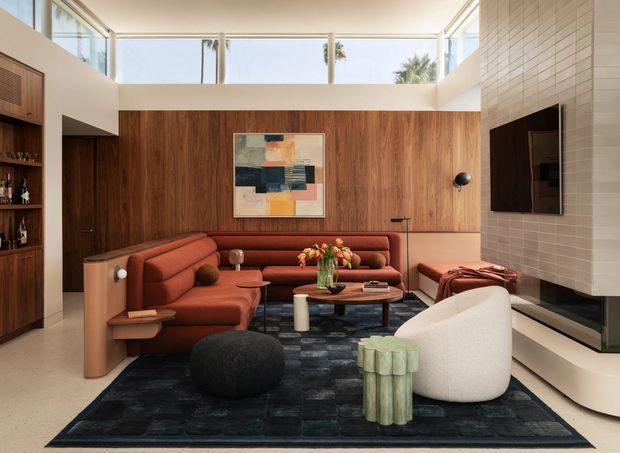 A sophisticated double-height living room with a warm walnut feature wall, integrated shelving and a wall-mounted television. A curved rust-coloured sectional sofa, sculptural white lounge chair, dark ottoman, coffee table and textured side table sit on a dark patterned rug. Clerestory glazing brings in abundant daylight, while a tiled fireplace wall and linear hearth occupy the right side.