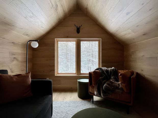 A compact, cozy attic-style living room with a steep gable ceiling clad in pale timber boards and warm timber walls. A central double window brings in filtered natural light, while a dark sofa, cognac leather armchair, draped throw, floor lamp, side table, and low rounded coffee table create a rustic retreat atmosphere. A small mounted deer head is positioned above the window.