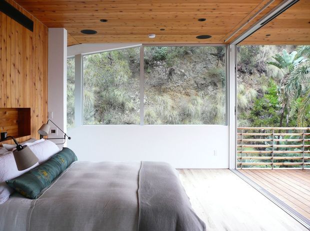 Bright bedroom with a built-in timber-paneled wall and matching timber ceiling, large floor-to-ceiling glazing, and an open connection to a timber deck. The room overlooks dense natural vegetation and has a low-profile bed with muted green and neutral bedding, a wall-mounted timber headboard, and a simple adjustable reading lamp. The architecture emphasizes indoor–outdoor connection and abundant natural light.