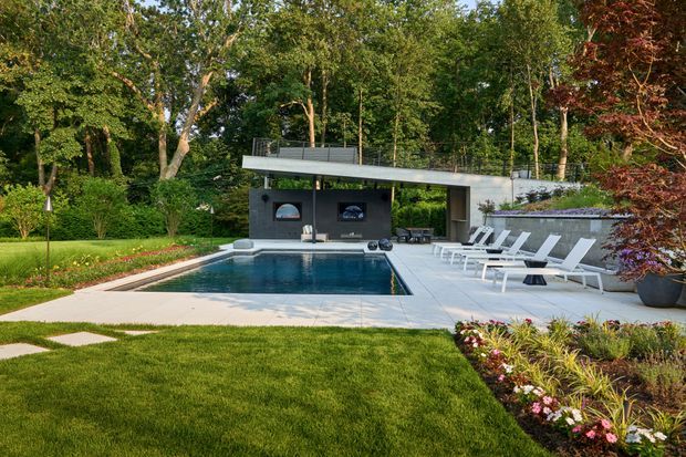A landscaped residential pool area in a wooded garden, featuring a long rectangular swimming pool, pale paved terrace, multiple white sun loungers, planting beds, and a compact modern pool pavilion with a flat roof, dark walls, outdoor kitchen or bar, and rooftop glass balustrade.