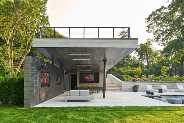 A modern concrete outdoor entertaining and pool area set within a landscaped garden. The structure features a broad flat-roofed cantilevered canopy, exposed concrete soffit, dark masonry walls, slender columns, integrated ceiling lighting, outdoor seating, and a sheltered media or lounge zone. Steps lead to an elevated garden and sports court area, while a paved terrace and pool edge occupy the foreground.
